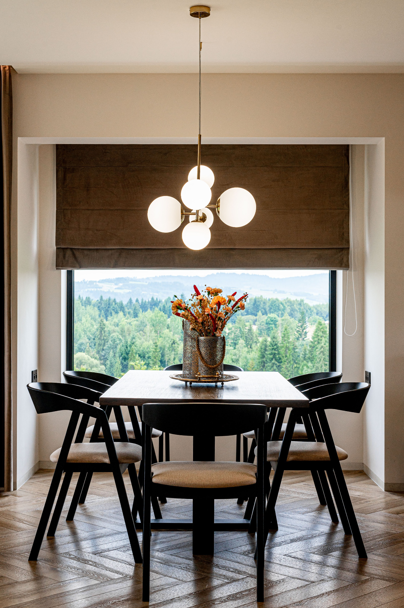 Stół z czarnymi krzesłami i bukietem kwiatów w wazonie na tle okna z widokiem na las. Nad stołem wisi nowoczesna lampa z kulistymi kloszami.