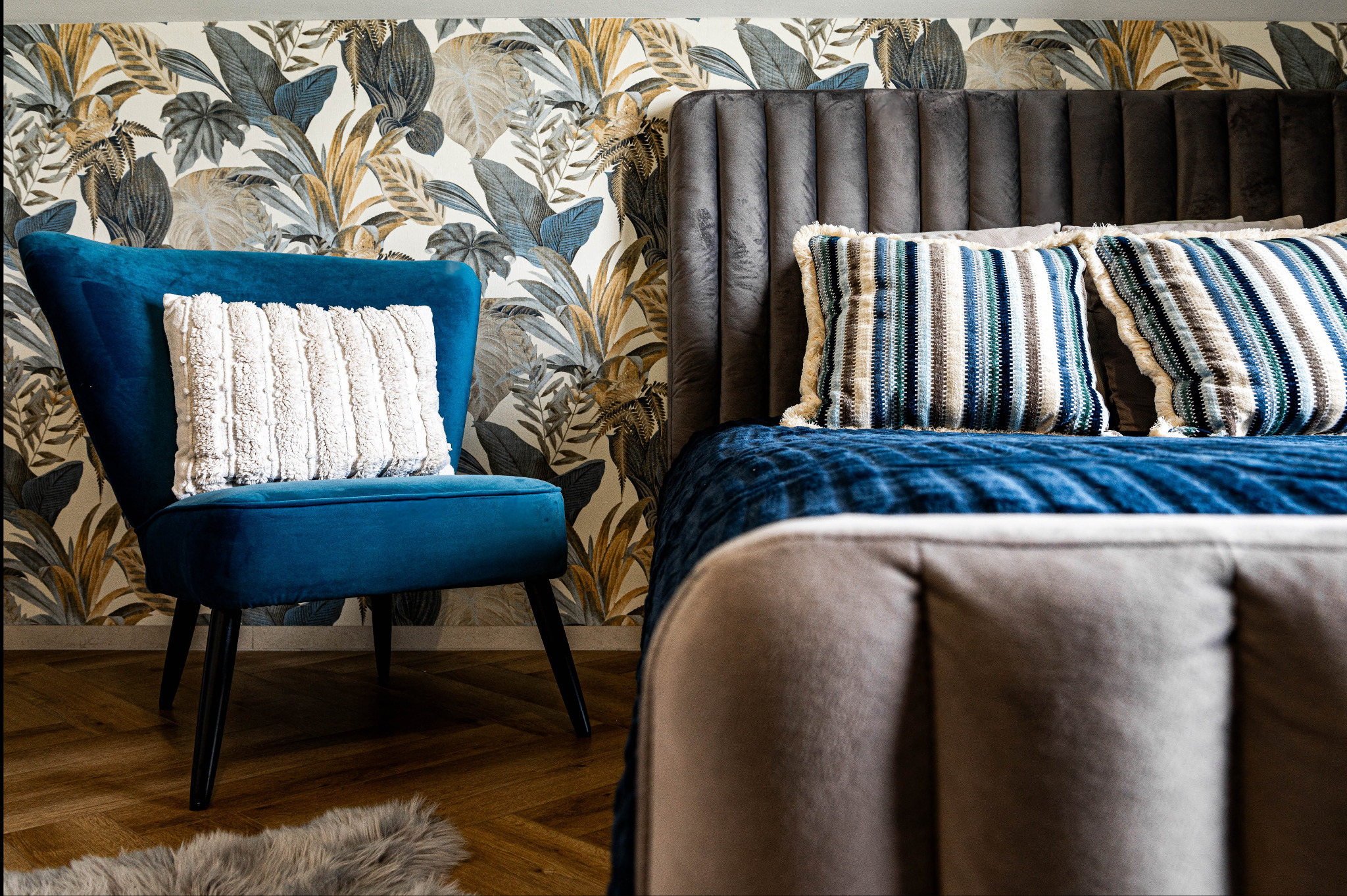 Stylowa sypialnia z turkusowym fotelem i wzorzystą tapetą w tropikalne liście. Szare łóżko z niebieską narzutą i dekoracyjnymi poduszkami.