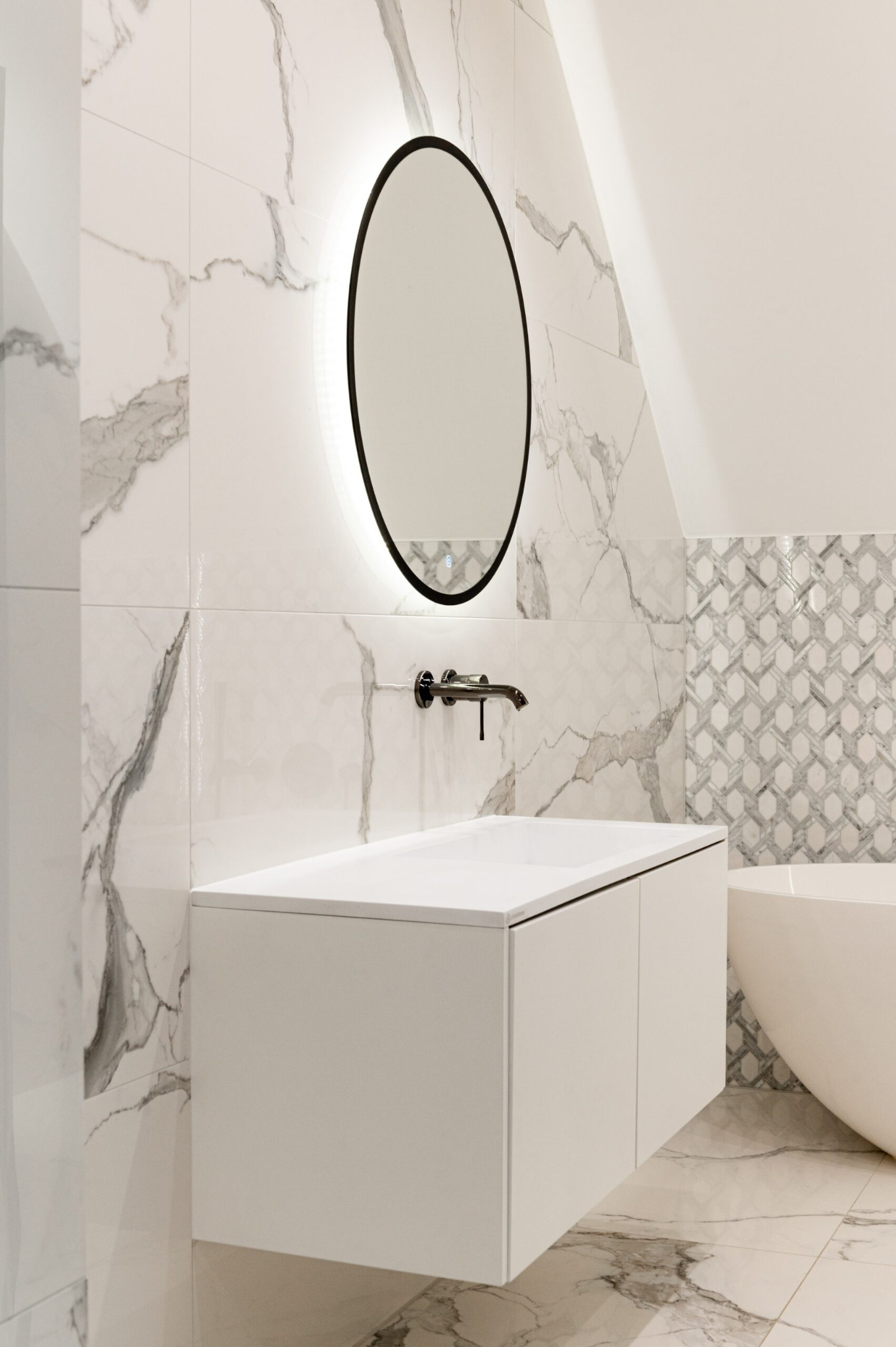 Nowoczesna łazienka w bieli z marmurowymi płytkami, okrągłym lustrem z podświetleniem i minimalistyczną szafką pod umywalką. Widoczny fragment wanny.
