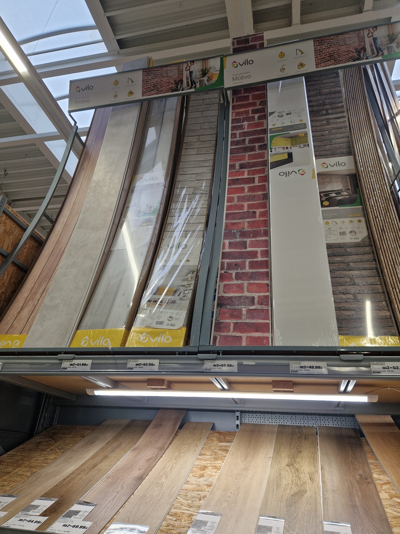 Panele dekoracyjne Vilo Motivo z wzorami cegły, drewna i kamienia na półce w sklepie budowlanym, oświetlone lampą. Wybór materiałów wykończeniowych.