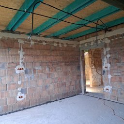 ELEKTROINSTALACJE KRZYSZTOF GRZYB - Wnętrze w trakcie prac: widoczne kable elektryczne prowadzone po suficie z desek i po ścianach z cegieł, z puszkami instalacyjnymi. Surowy stan, brak wykończenia.