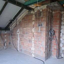 ELEKTROINSTALACJE KRZYSZTOF GRZYB - Wnętrze w trakcie prac wykończeniowych: murowana ściana z widocznymi przewodami elektrycznymi, częściowo zamaskowanymi gipsem. Surowy stan, brak tynków.