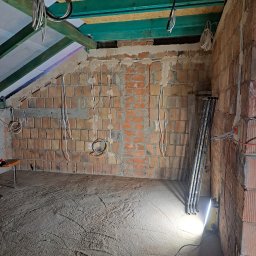 ELEKTROINSTALACJE KRZYSZTOF GRZYB - Surowy stan poddasza z instalacją elektryczną w bruzdach ceglanej ściany, widoczne kable zwisające z sufitu i prowizoryczne oświetlenie budowlane.
