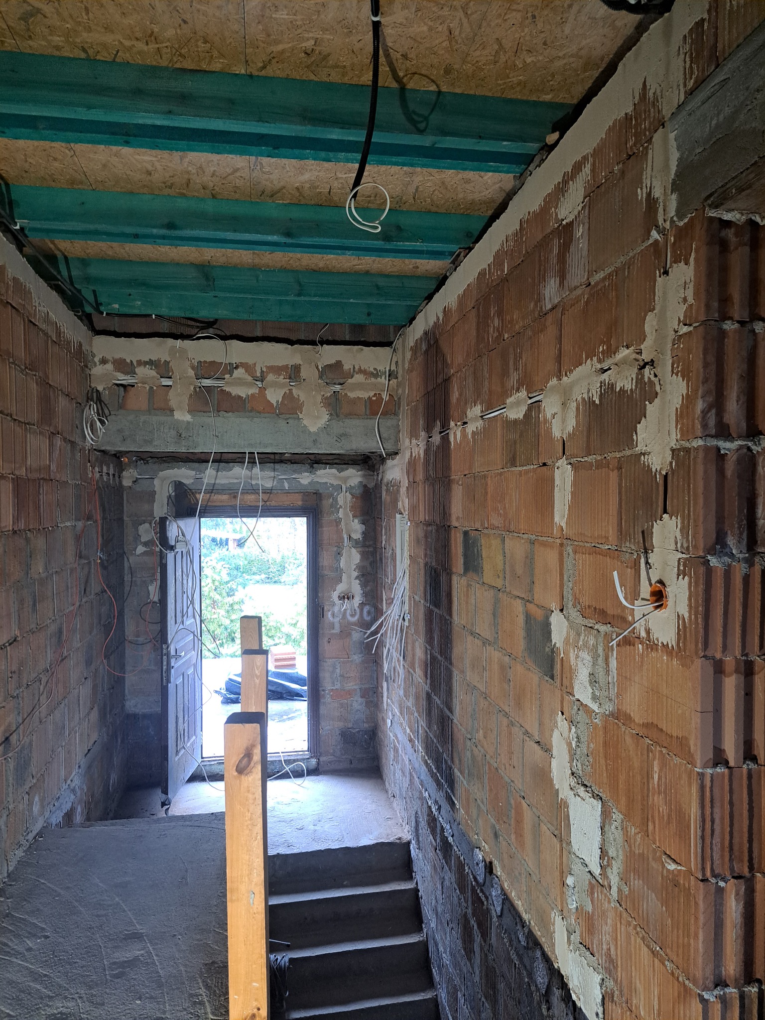 Surowy stan klatki schodowej z widoczną instalacją elektryczną w ścianach z czerwonej cegły i drewnianym stropem z zielonymi belkami. Otwarta brama w tle.