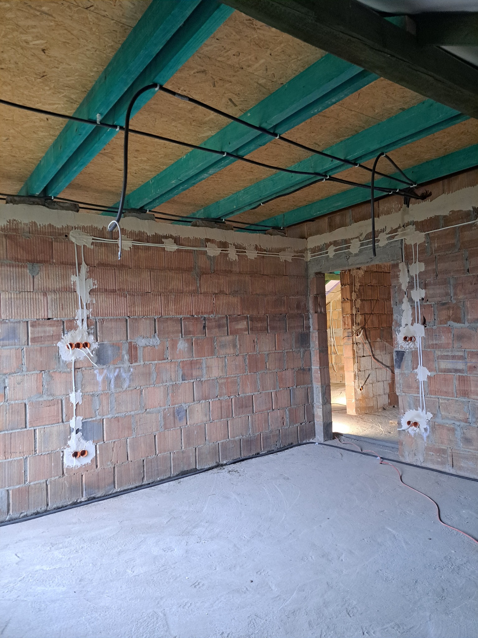 Wnętrze w trakcie prac: widoczne kable elektryczne prowadzone po suficie z desek i po ścianach z cegieł, z puszkami instalacyjnymi. Surowy stan, brak wykończenia.