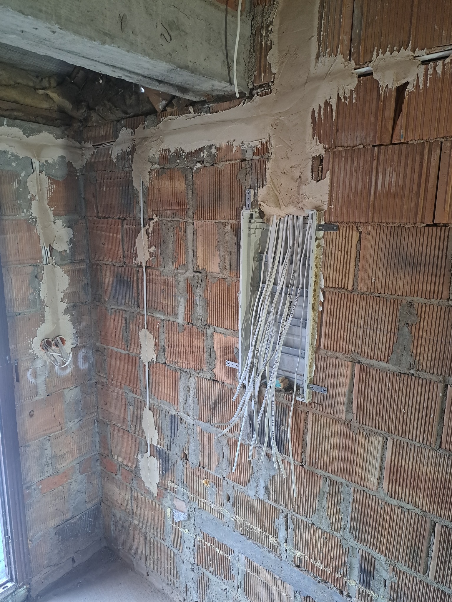Montaż skrzynki elektrycznej w surowym murze z cegły. Widoczne przewody i zaprawa. Instalacja elektryczna w trakcie realizacji.