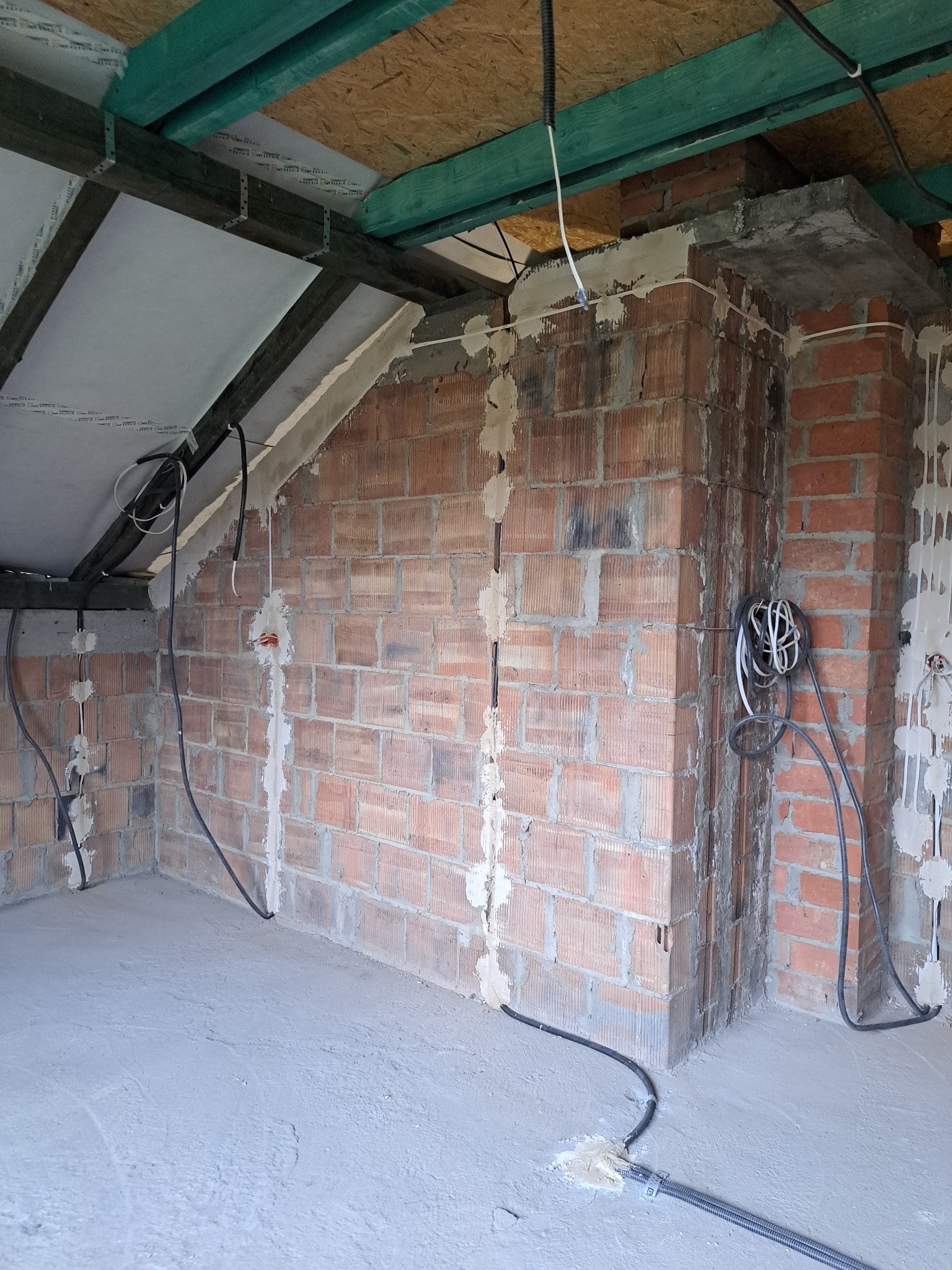 Wnętrze w trakcie prac wykończeniowych: murowana ściana z widocznymi przewodami elektrycznymi, częściowo zamaskowanymi gipsem. Surowy stan, brak tynków.