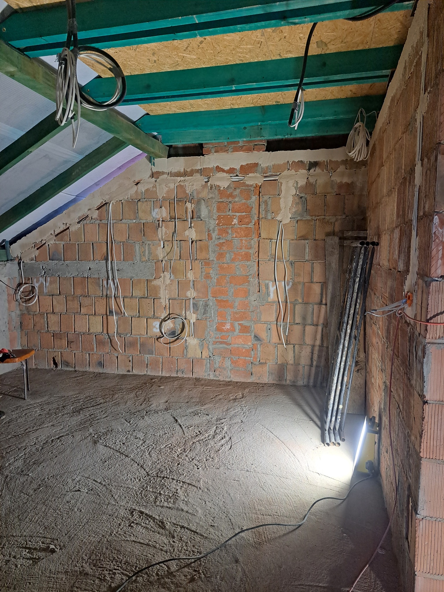 Surowy stan poddasza z instalacją elektryczną w bruzdach ceglanej ściany, widoczne kable zwisające z sufitu i prowizoryczne oświetlenie budowlane.
