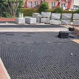 Usługi ziemne oraz ogrodnicze - Układanie ekokratki na podjeździe. Pracownik zagęszcza podłoże ubijarką. W tle kostka brukowa i palety z materiałem. Widoczne narzędzia i butelki z wodą.