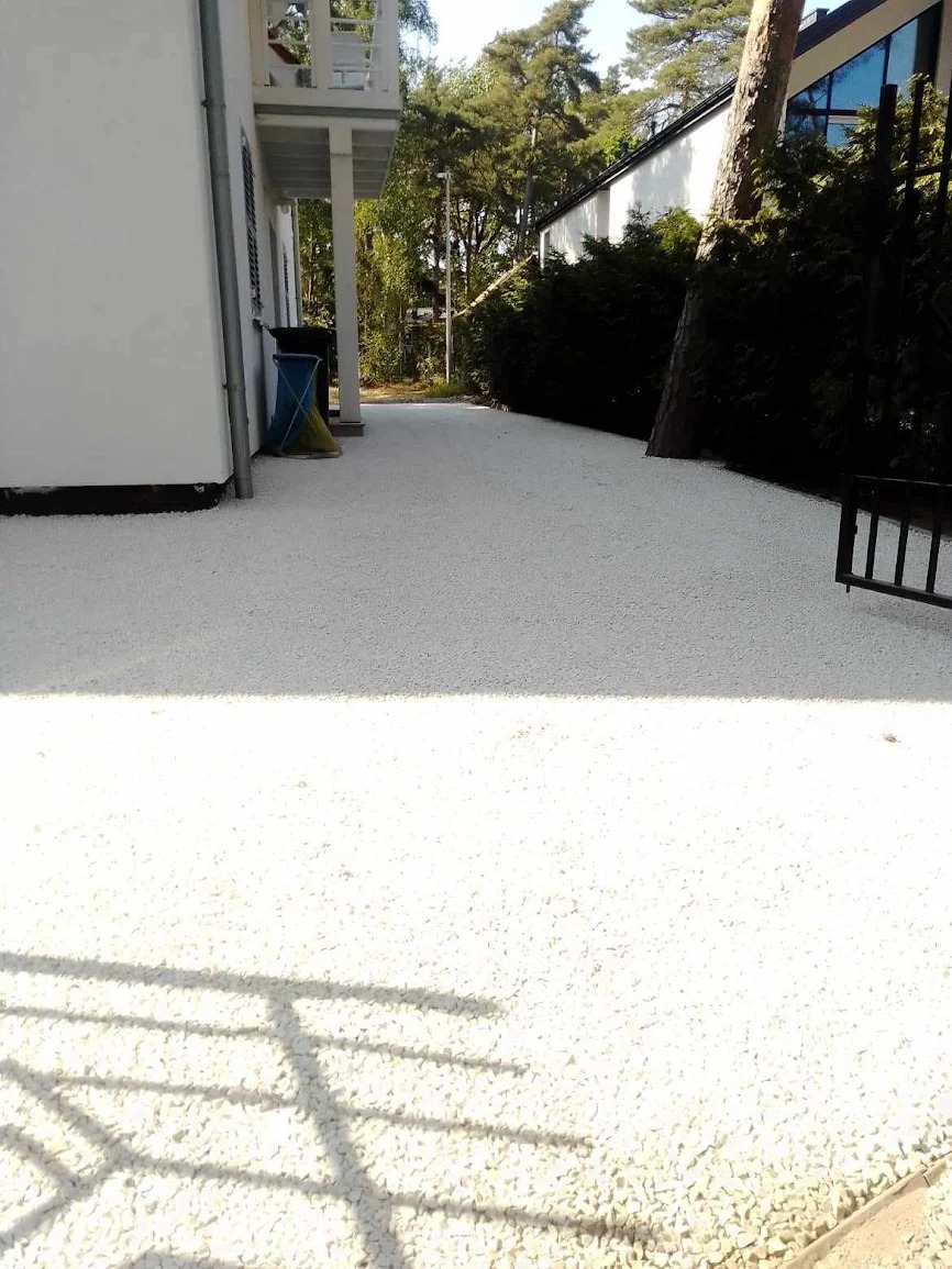 Biała, kamienna nawierzchnia przed budynkiem z białymi ścianami i zielenią w tle. Widoczny fragment balustrady i drzewo. Jasne, słoneczne oświetlenie.