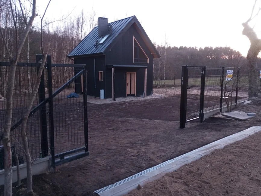 Nowoczesny, czarny dom z metalowym dachem i drewnianymi drzwiami, widziany zza otwartej, metalowej bramy w surowym ogrodzie z widocznym systemem odwadniającym.