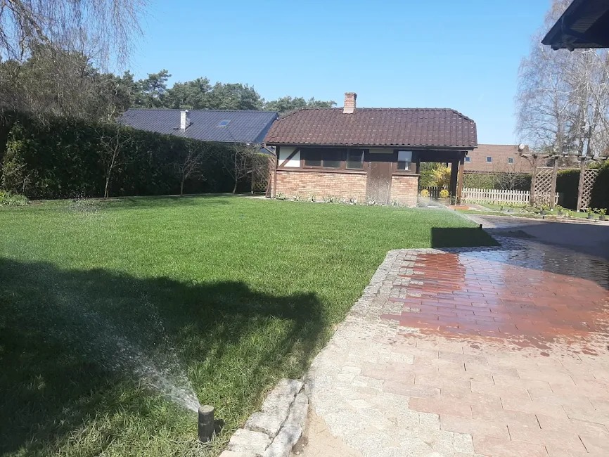 Zraszacz nawadnia soczysty trawnik w Gryficach. W tle budynek z cegły i zadbany ogród. Widoczny fragment utwardzonej drogi z kostki brukowej.