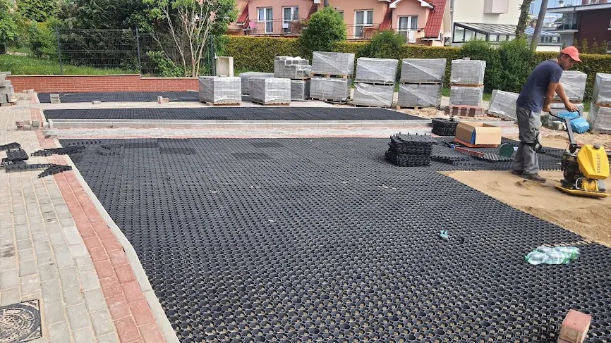 Układanie ekokratki na podjeździe. Pracownik zagęszcza podłoże ubijarką. W tle kostka brukowa i palety z materiałem. Widoczne narzędzia i butelki z wodą.