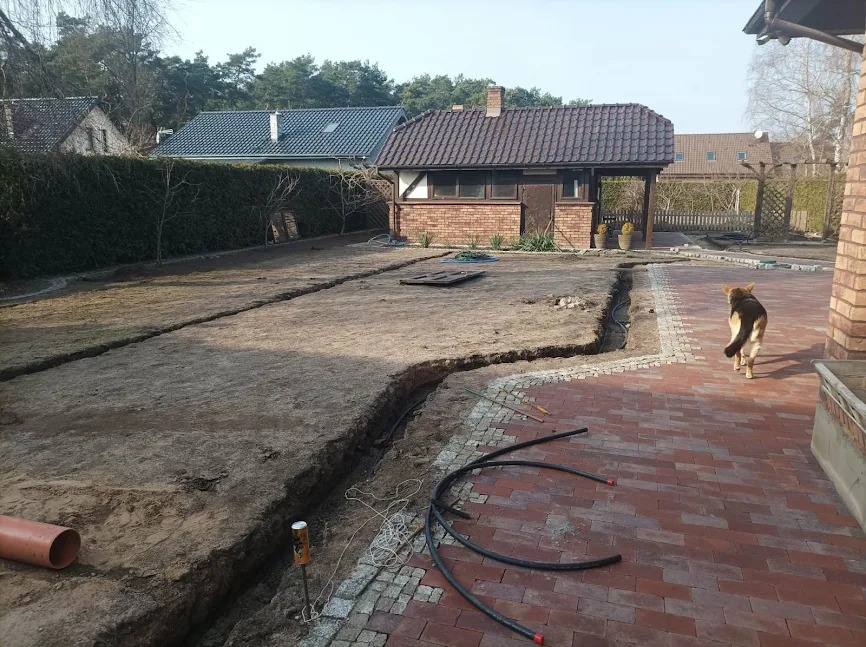 Wykopy w ziemi pod instalacje w ogrodzie, widoczne rury i kabel. Częściowo ułożona kostka brukowa z obrzeżami. Pies spaceruje po kostce. Dom w tle.