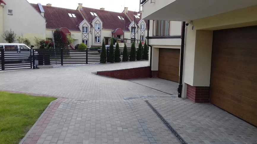 Jasnoszara kostka brukowa na podjeździe do garażu, z widoczną linią odpływową. W tle rząd tuj i brama wjazdowa z metalowymi przęsłami, domy w zabudowie szeregowej.