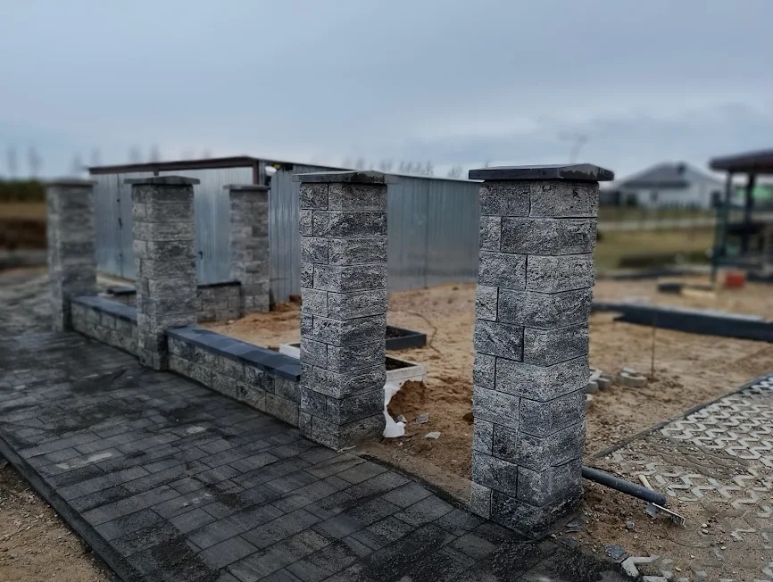 Szara kostka brukowa tworząca ścieżkę i słupki ogrodzeniowe z elementami dekoracyjnymi, w tle blaszany garaż i domy. Praca w toku, widoczne niedokończone elementy.