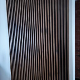 Top build-Wykończenie wnętrz - Drewniana ścianka działowa z pionowych listew w odcieniach brązu, oddzielająca białą ścianę od ściany z czerwonej cegły. Nowoczesny design wnętrza.