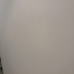 Top build-Wykończenie wnętrz - Gładka, pomalowana na jasny odcień szarości ściana w rogu pomieszczenia, z fragmentem podłogi w dolnej części kadru. Widoczne ślady po malowaniu.