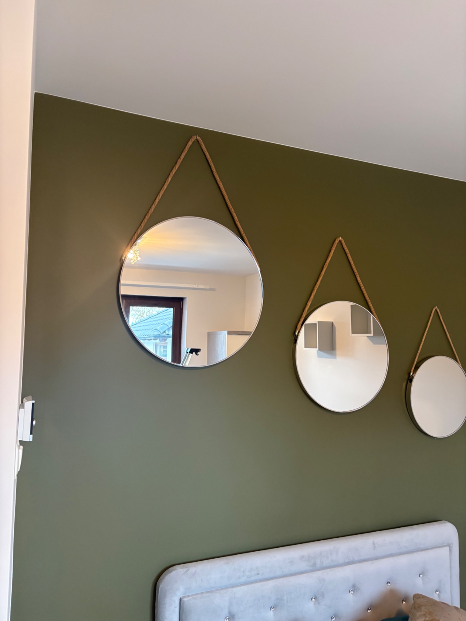 Oliwkowa ściana z trzema okrągłymi lustrami zawieszonymi na sznurkach, odbijającymi wnętrze pokoju. Widoczny fragment zagłówka łóżka.