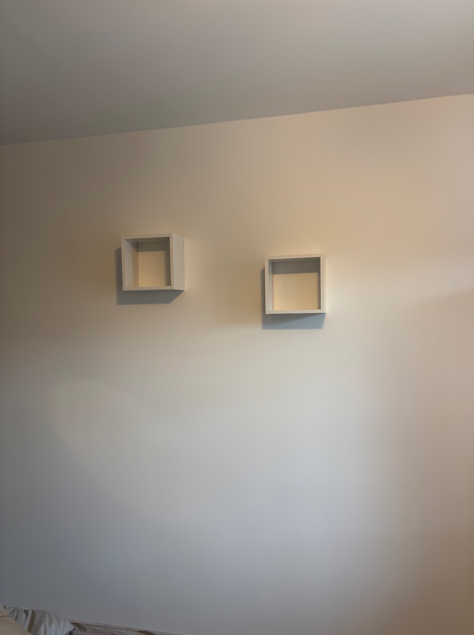 Dwie białe, kwadratowe półki zamontowane na kremowej ścianie. Półki są puste i mają minimalistyczny design. Oświetlenie pada z prawej strony, tworząc delikatny cień.