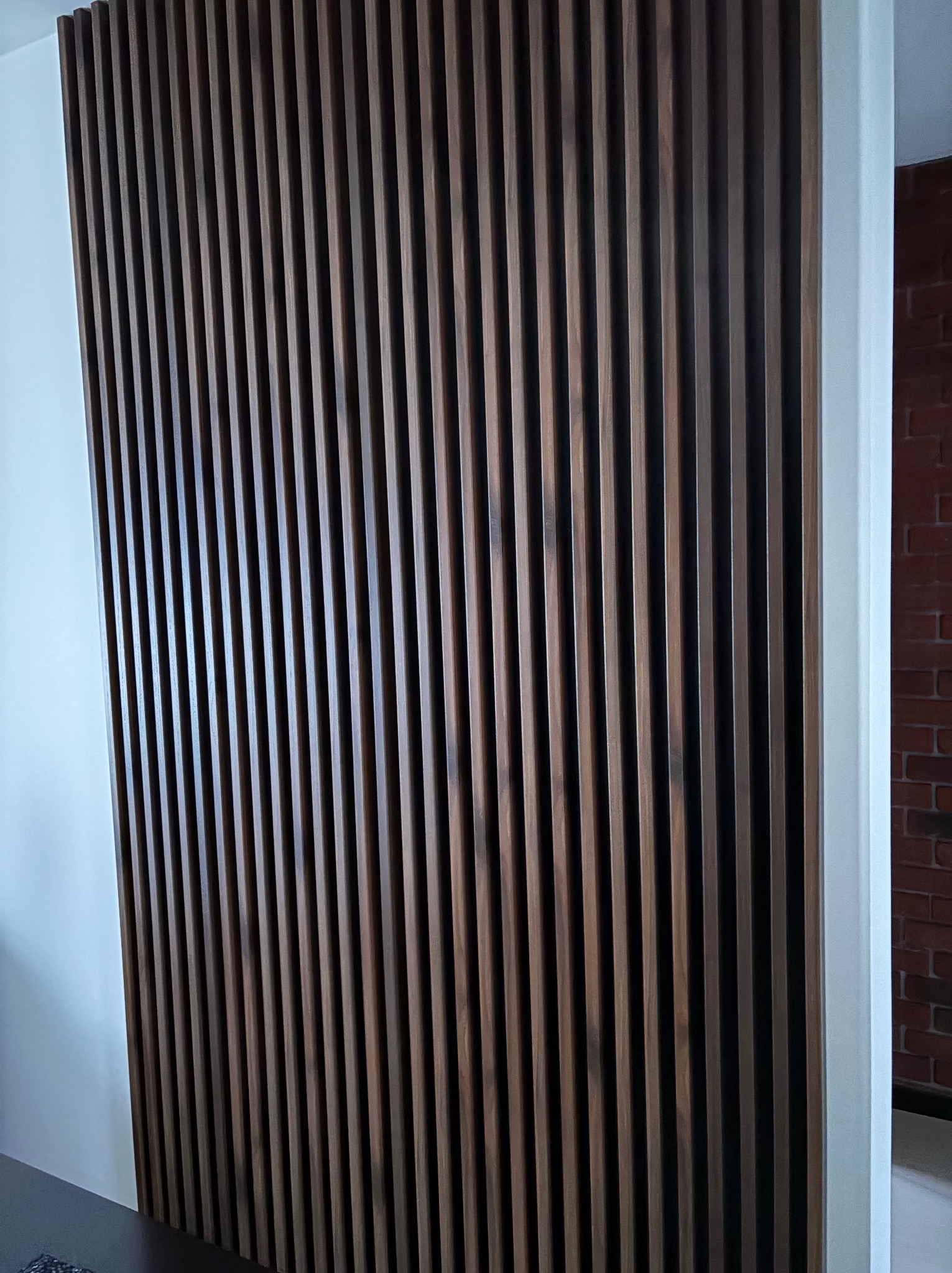 Drewniana ścianka działowa z pionowych listew w odcieniach brązu, oddzielająca białą ścianę od ściany z czerwonej cegły. Nowoczesny design wnętrza.