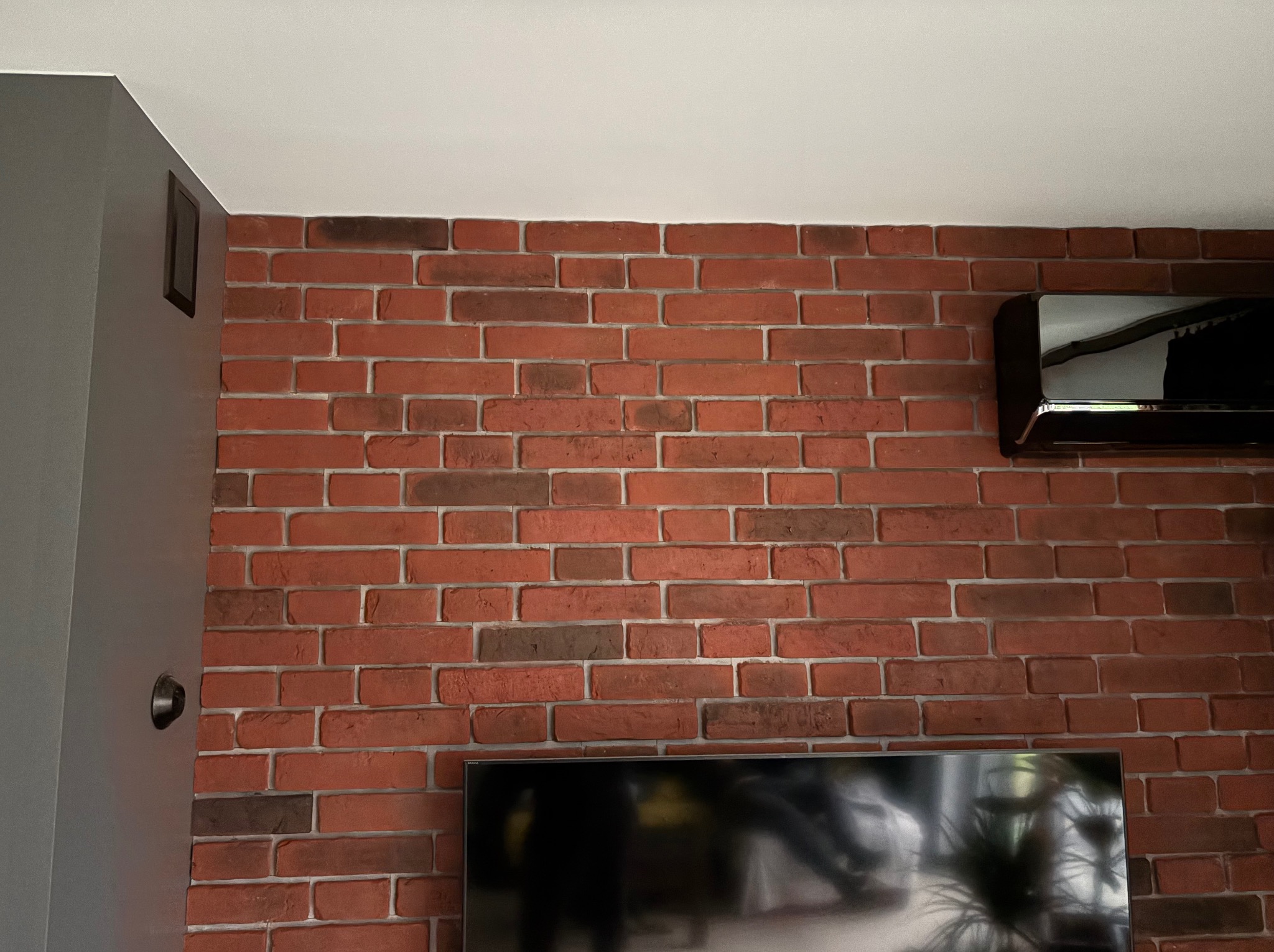 Ściana z cegły klinkierowej w salonie, z widocznym telewizorem i klimatyzatorem. Dekoracyjne wykończenie wnętrza z elementami szarości i bieli.