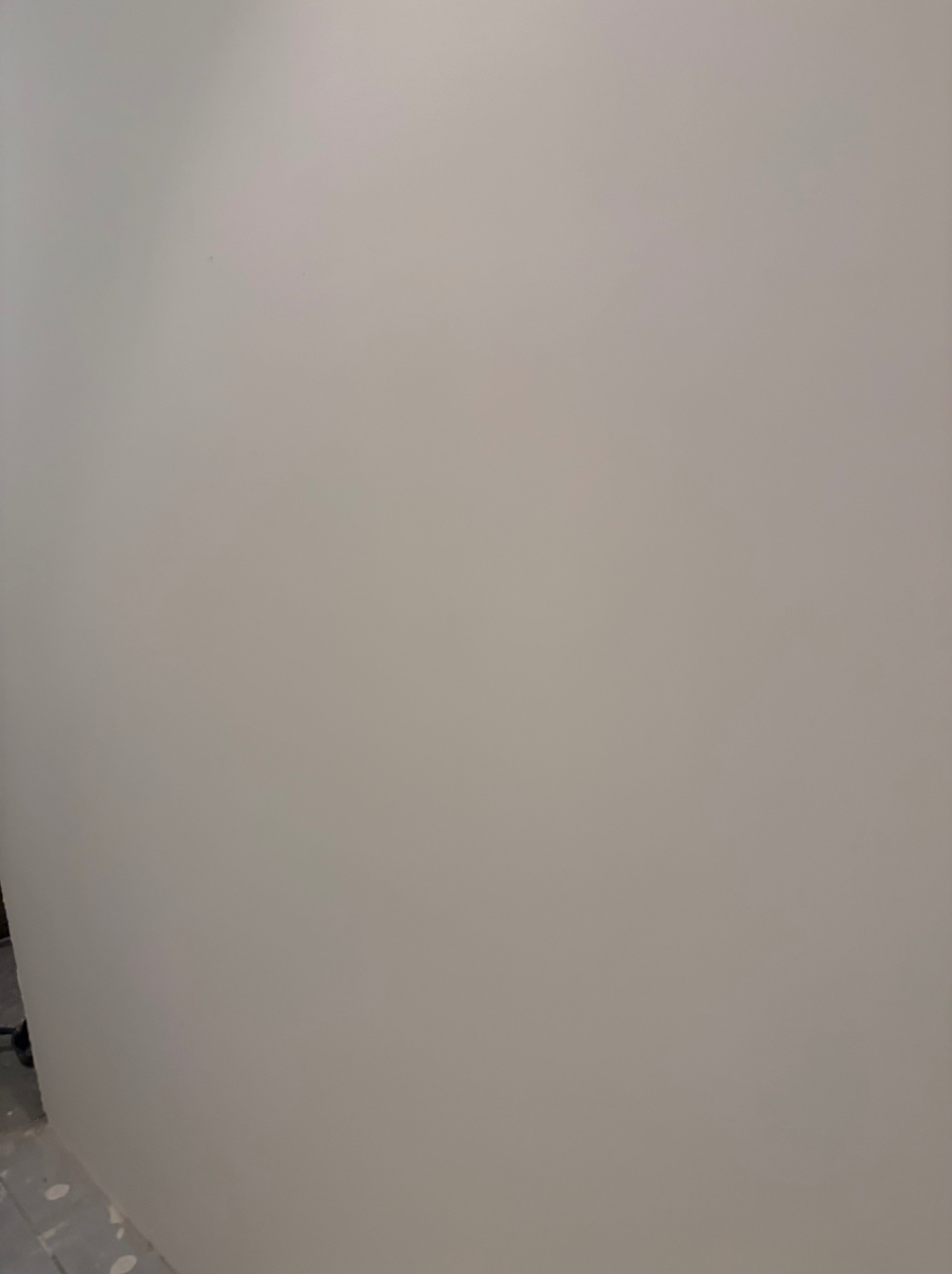 Gładka, pomalowana na jasny odcień szarości ściana w rogu pomieszczenia, z fragmentem podłogi w dolnej części kadru. Widoczne ślady po malowaniu.