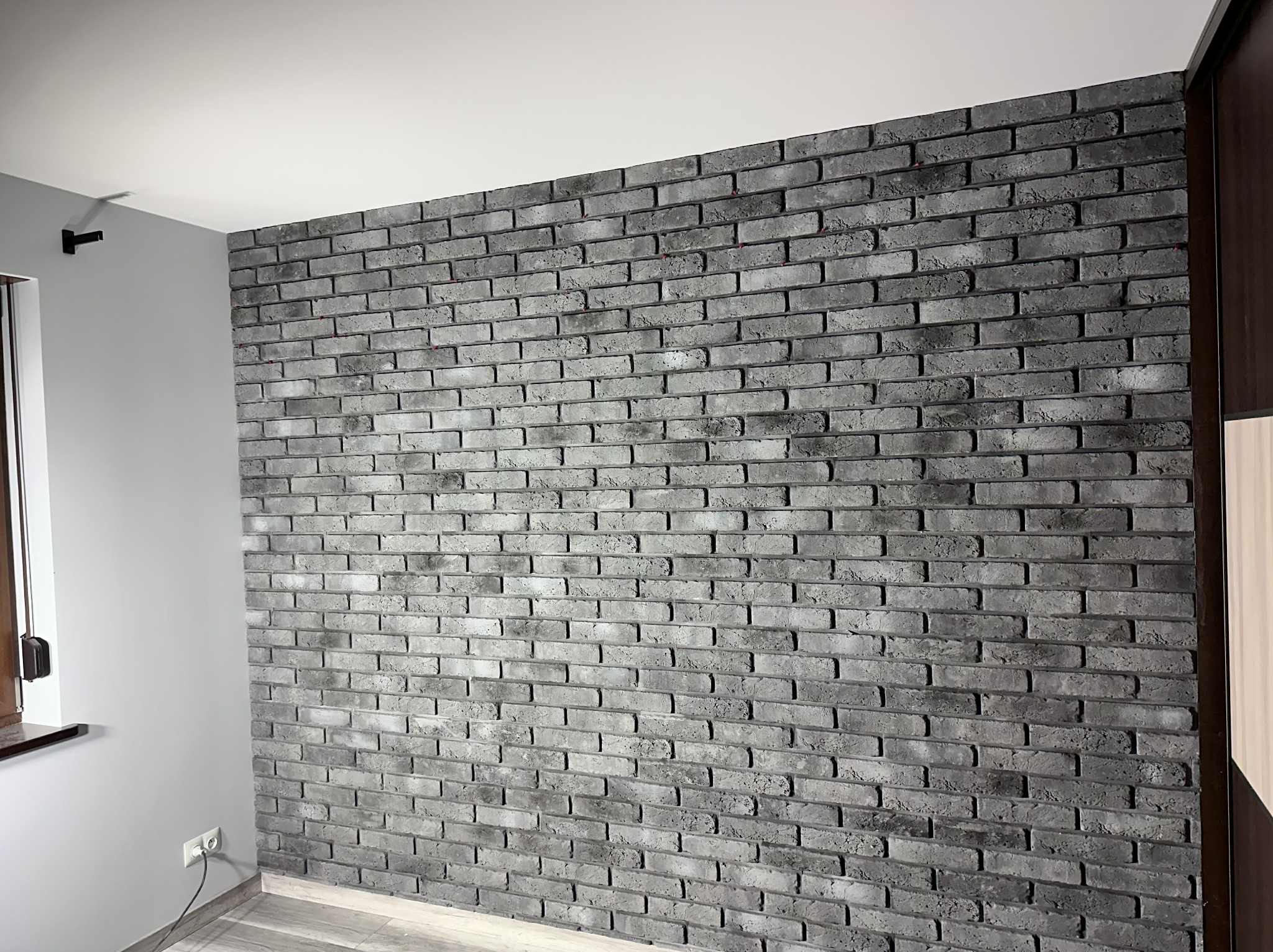 Ściana z dekoracyjnej szarej cegły w Grębocicach, z widocznym gniazdkiem elektrycznym i fragmentem okna po lewej stronie. Minimalistyczny design wnętrza.