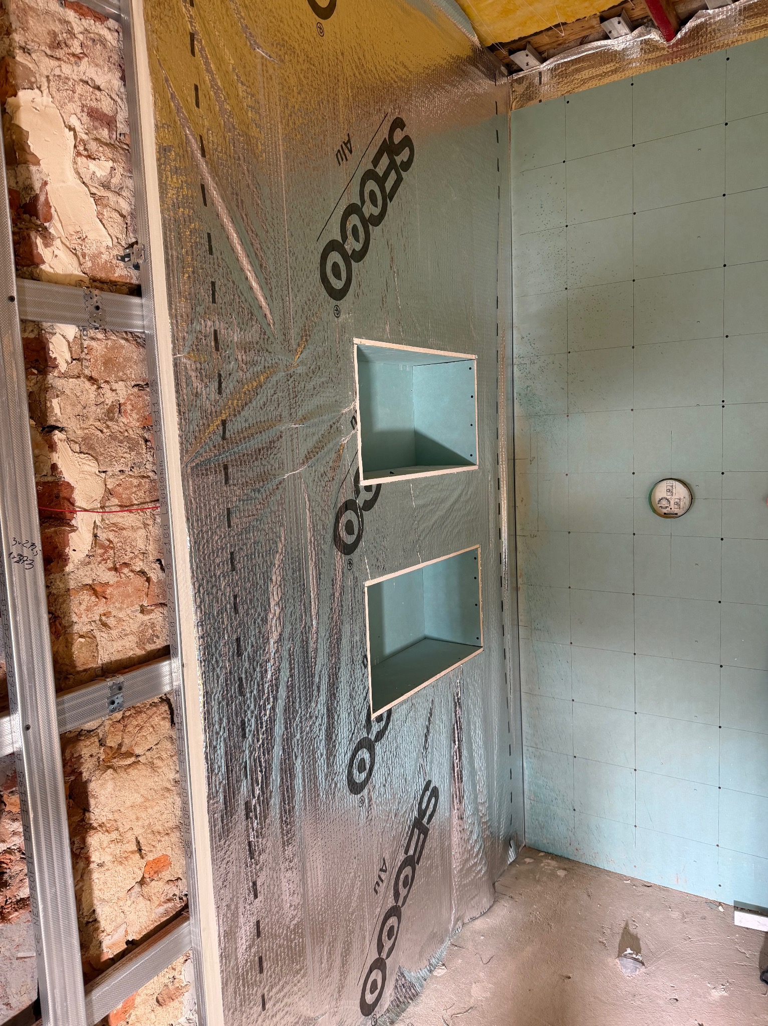 Łazienka w trakcie remontu: widoczne stelaże, izolacja z folii aluminiowej, płyty gipsowo-kartonowe z zaznaczonymi liniami i wnęki na półki. Surowy stan, przygotowanie do dalszych prac.