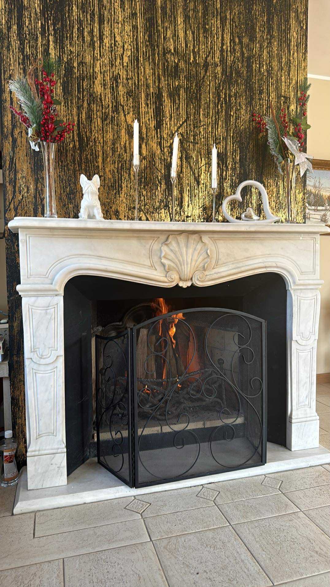 Marmurowy kominek z paleniskiem za ozdobną kratą, udekorowany świecami, figurką psa i kwiatami na tle ściany z efektem złocenia.