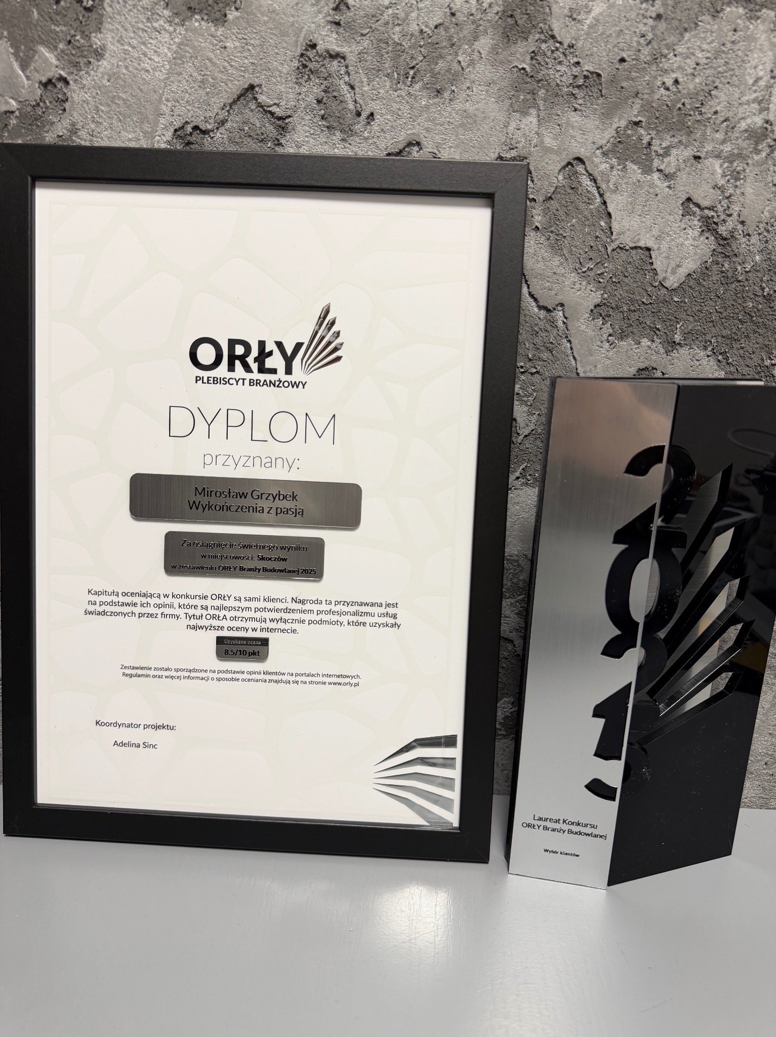 Dyplom 'Orły Branży Budowlanej' dla Mirosława Grzybka z 'Wykończenia z pasją' oraz statuetka laureata na tle szarej, teksturowanej ściany. Nagroda za opinie klientów.
