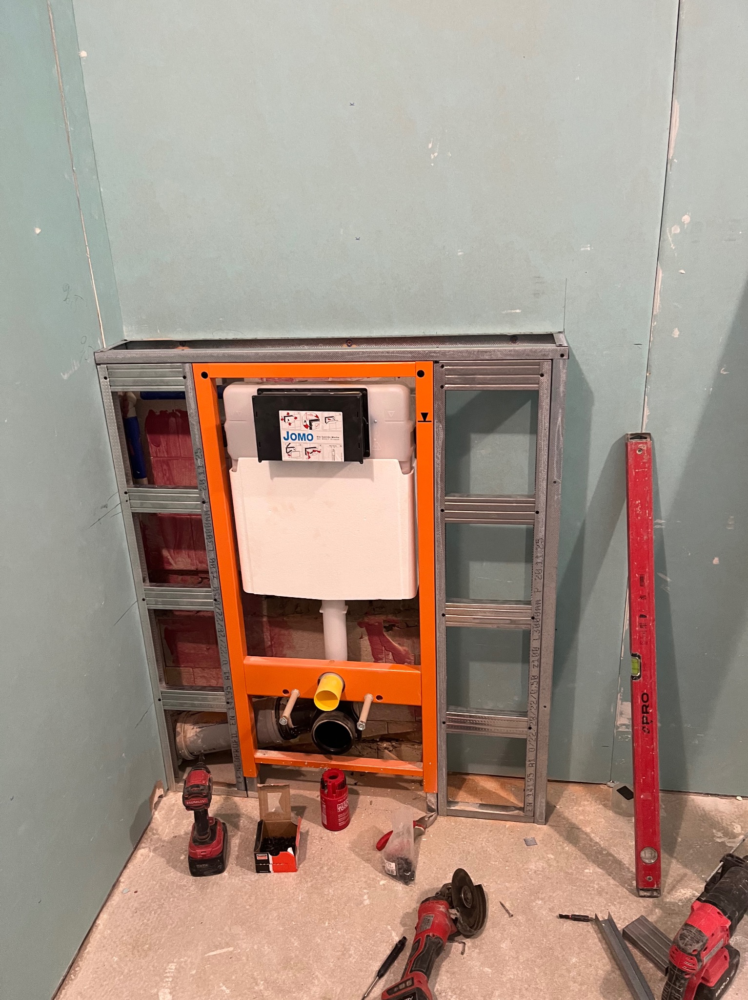 Stelaż podtynkowy WC w trakcie montażu, widoczne narzędzia i poziomica. Pomieszczenie w surowym stanie, ściany z płyt kartonowo-gipsowych.