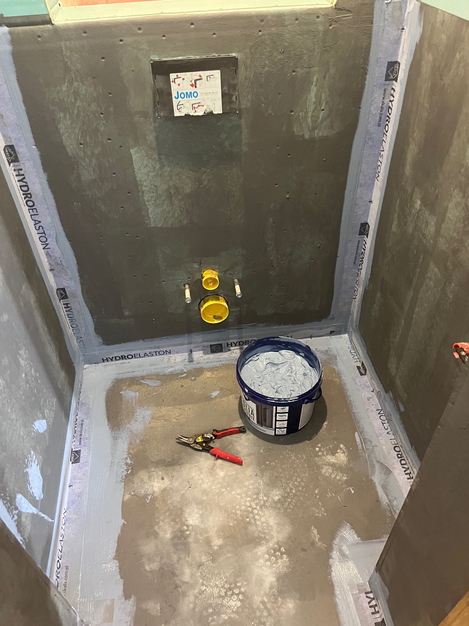 Hydroizolacja w kabinie prysznicowej w trakcie prac. Widoczna puszka JOMO, wiadro z masą uszczelniającą i narzędzia. Hydroelaston na taśmach uszczelniających.