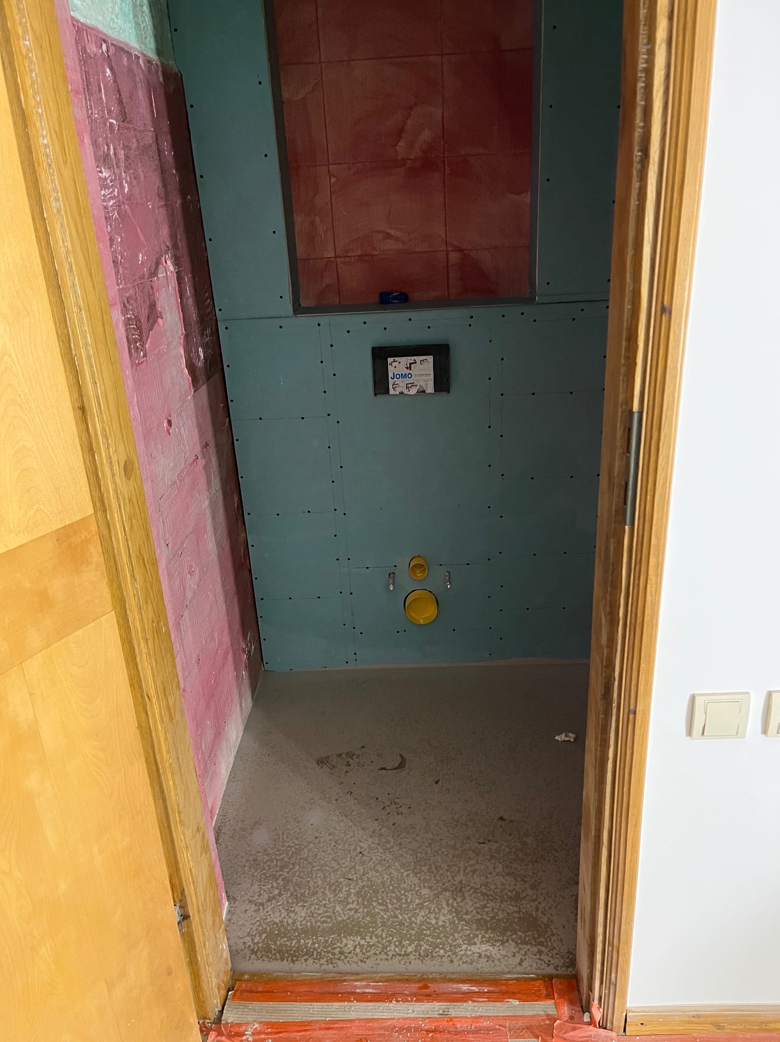 Łazienka w trakcie remontu: ściany z płyt gipsowo-kartonowych, widoczne instalacje, przygotowana podłoga. Ujęcie zza futryny drzwi.