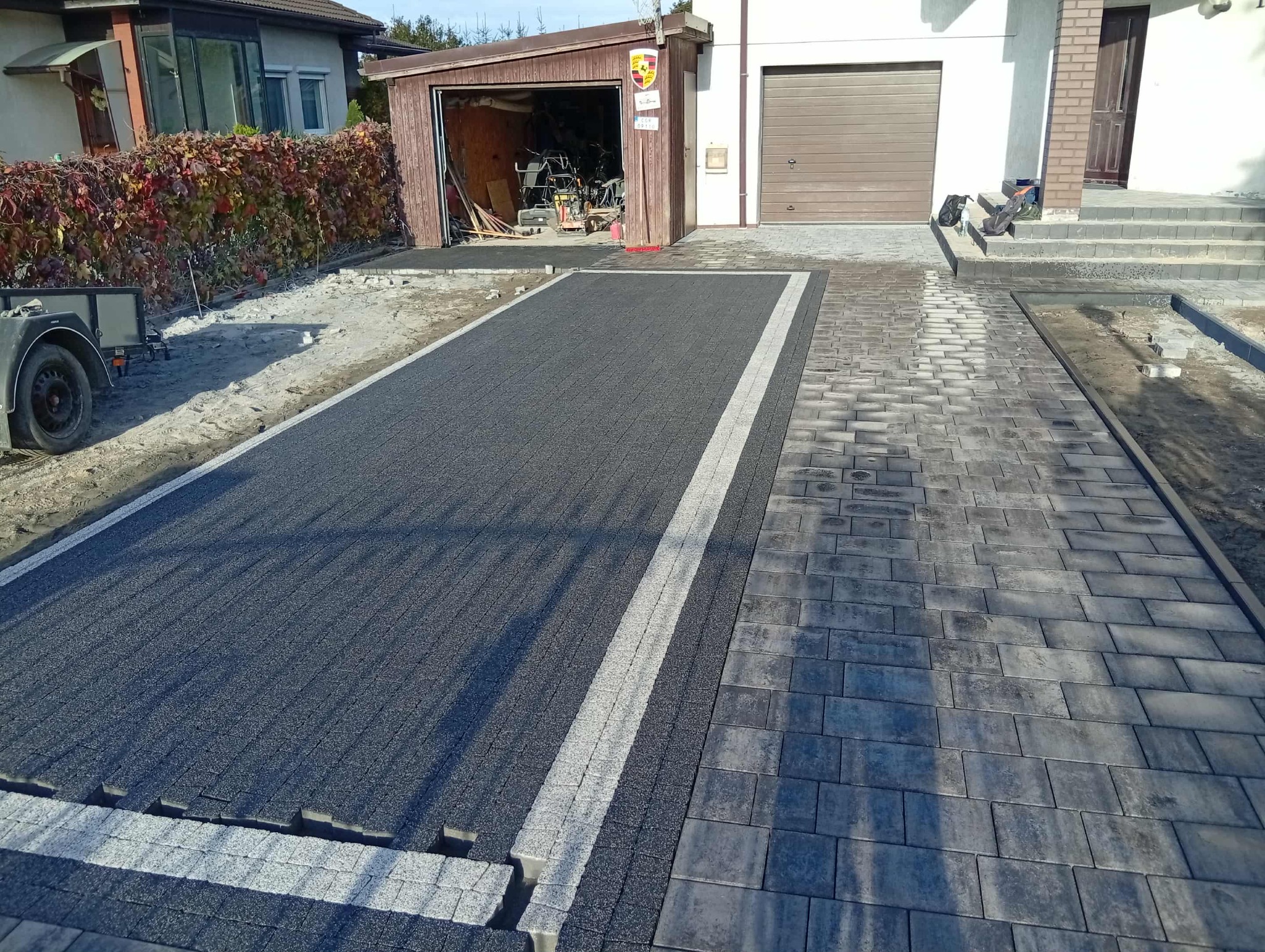 Ułożona kostka brukowa w dwóch odcieniach szarości, tworząca podjazd i ścieżkę. Widoczny garaż z otwartymi drzwiami i narzędzia. Częściowo ułożona kostka.