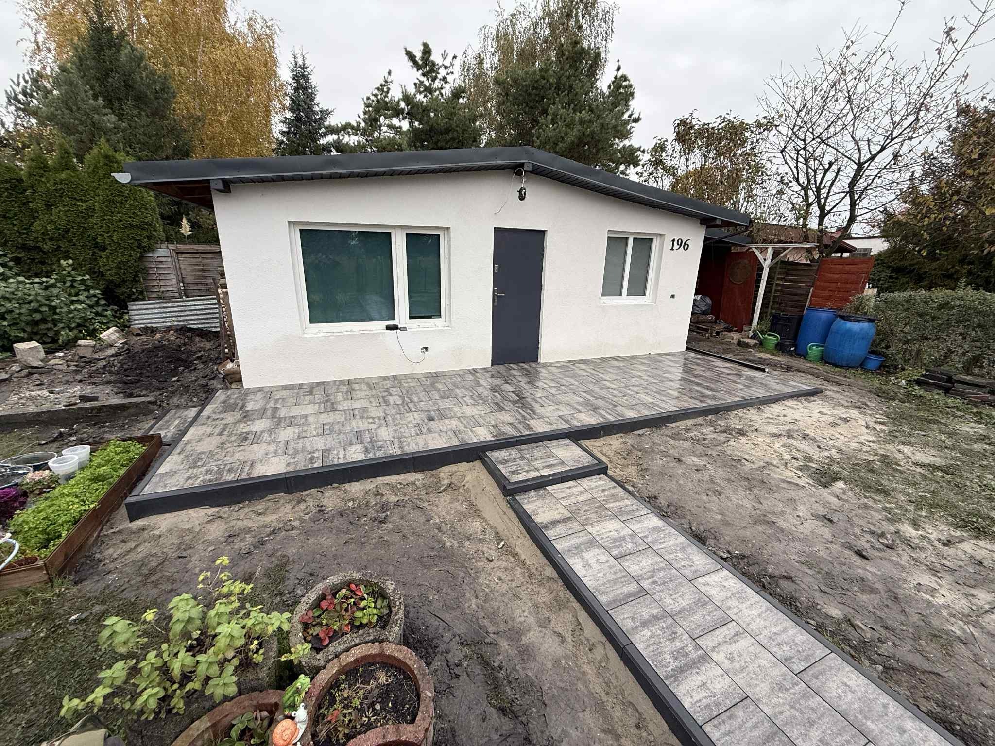 Nowoczesny taras z szarej kostki brukowej przylegający do białego budynku z ciemnymi oknami i drzwiami. Widoczne donice z roślinami i fragment ogrodu.