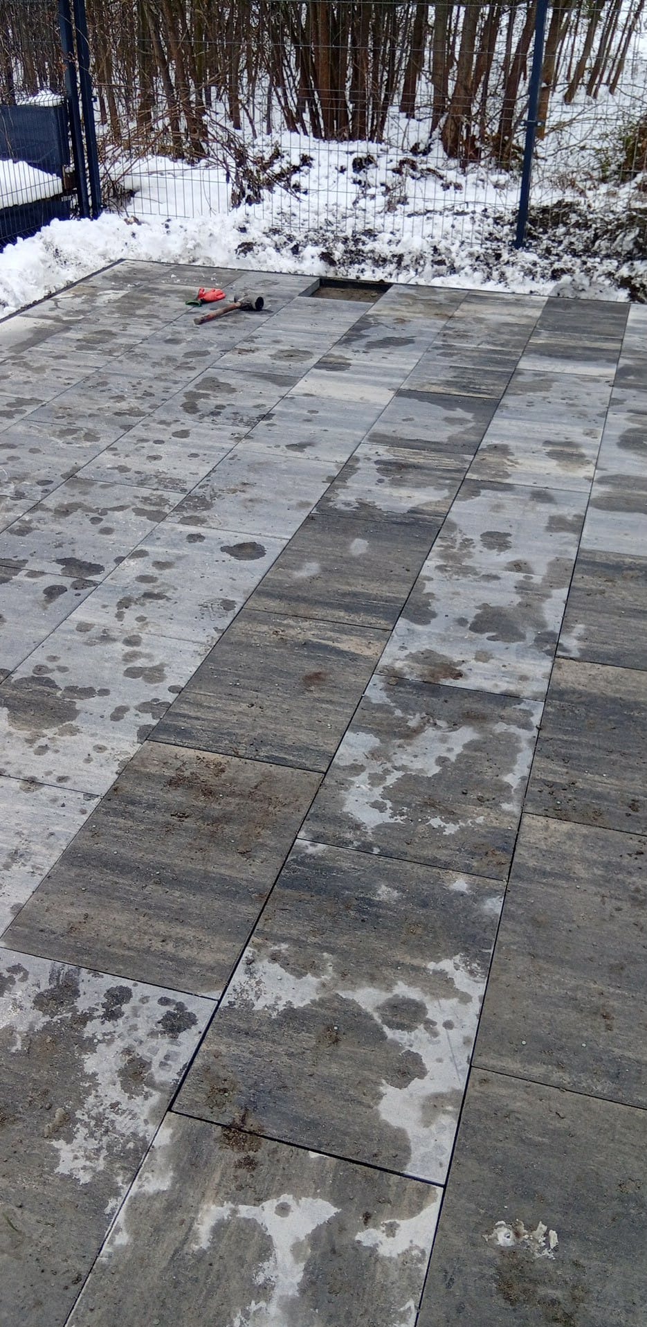 Ułożona kostka brukowa na zewnątrz w zimowej scenerii. Widoczne narzędzia: młotek i rękawice. Część kostki jeszcze nieułożona.