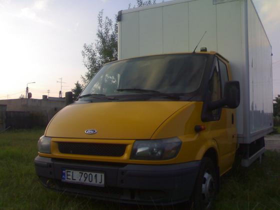 Żółty samochód dostawczy z białą zabudową kontenerową na trawniku, widoczne tablice rejestracyjne i budynek w tle.