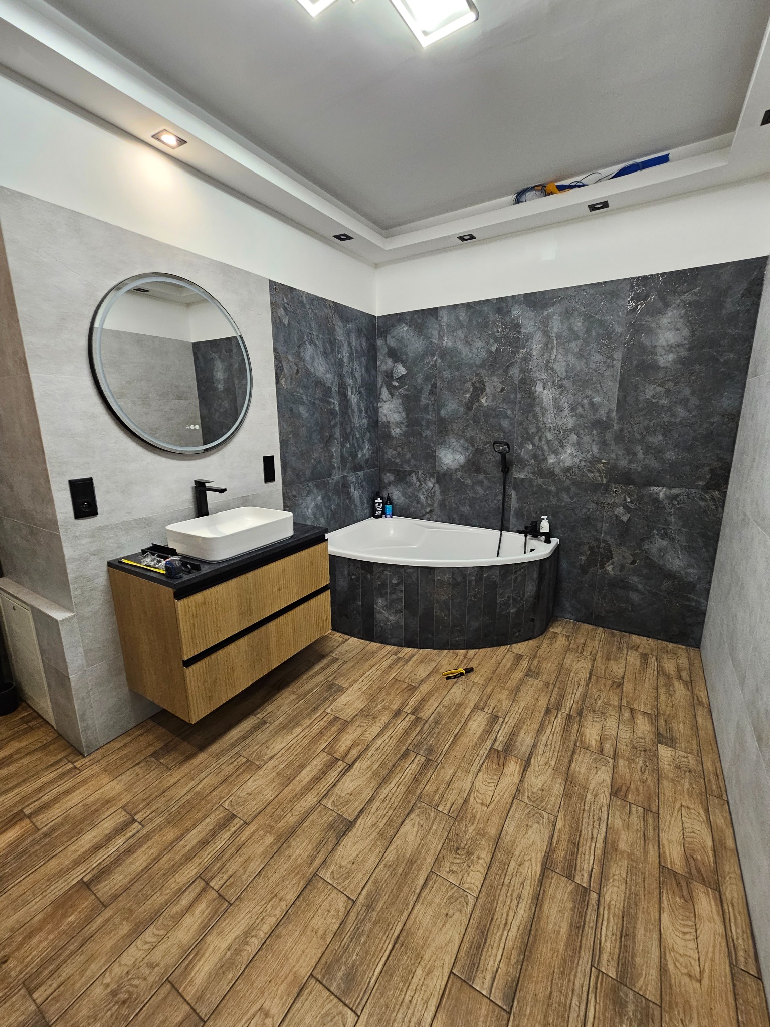 Nowoczesna łazienka po remoncie: drewnopodobna podłoga, grafitowe płytki, owalne lustro z podświetleniem i biała wanna narożna. Widoczne narzędzia.