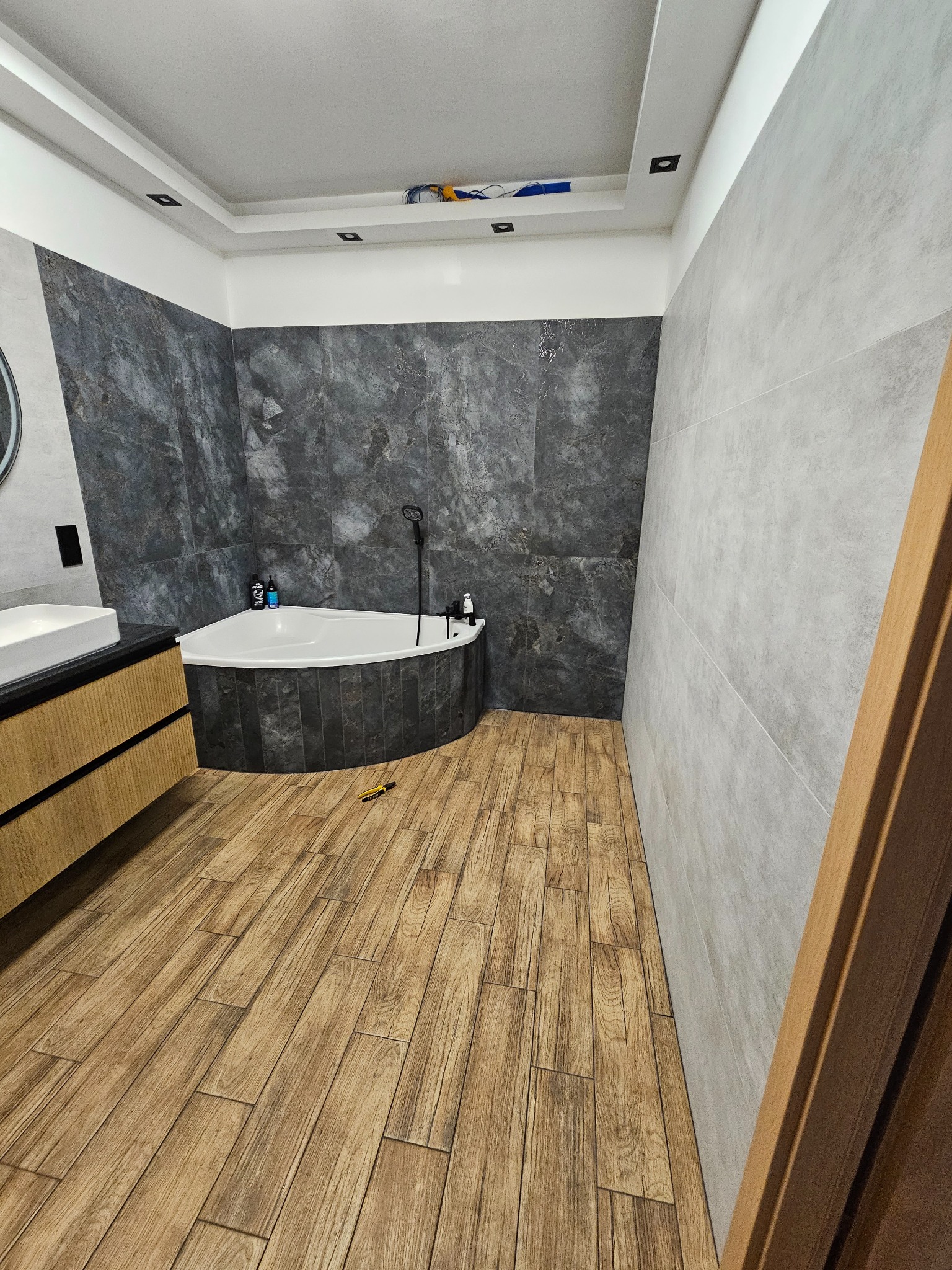 Nowoczesna łazienka po remoncie: narożna wanna obudowana ciemnymi płytkami, drewnopodobna podłoga, szare ściany. Widoczne narzędzia na podłodze.