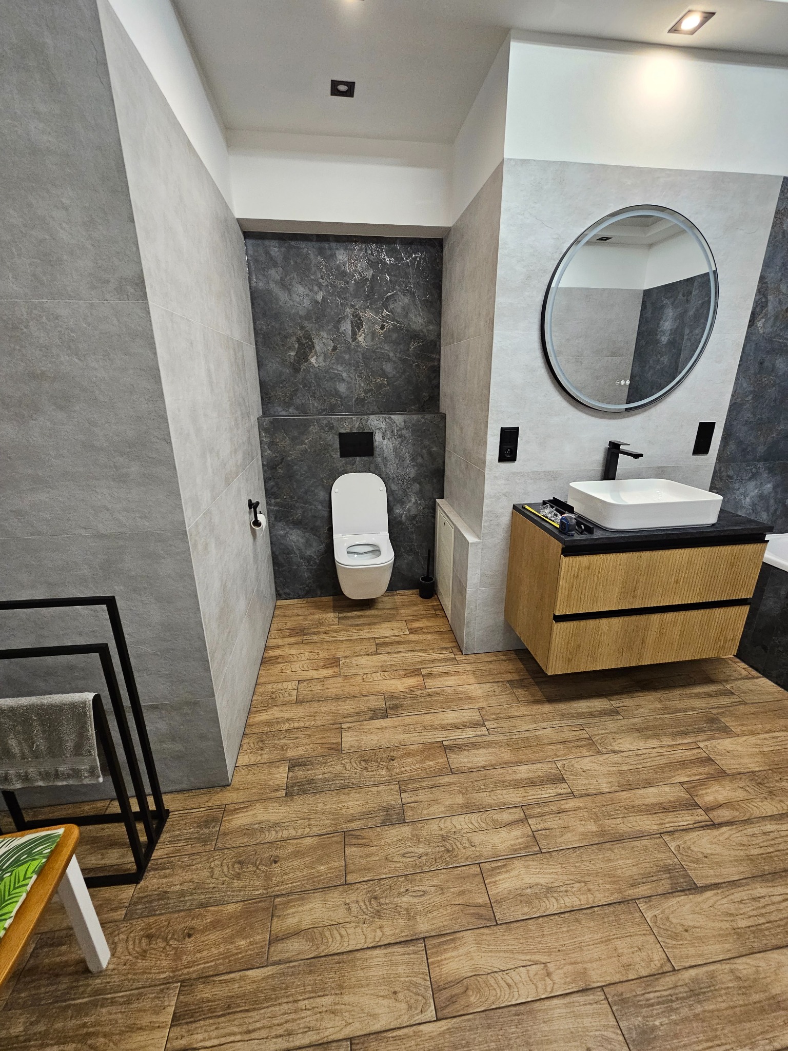 Nowoczesna łazienka z drewnopodobną podłogą, szarą glazurą i okrągłym lustrem. Umywalka nablatowa na szafce z drewnianymi frontami. Toaleta podwieszana.