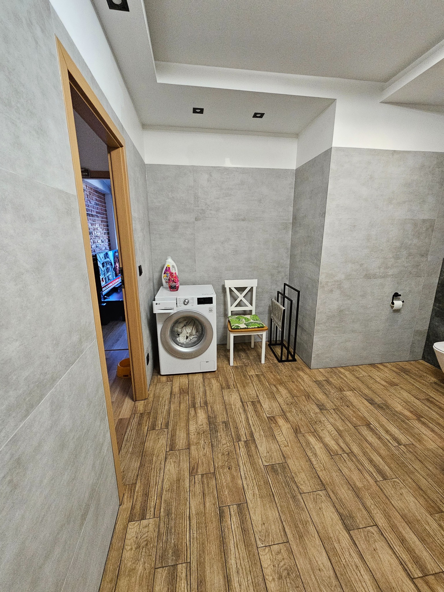 Wykończona łazienka z drewnopodobną podłogą, szarą glazurą i pralką. Widok na wnętrze przez otwarte drzwi z drewnianą ościeżnicą. Minimalistyczny design.