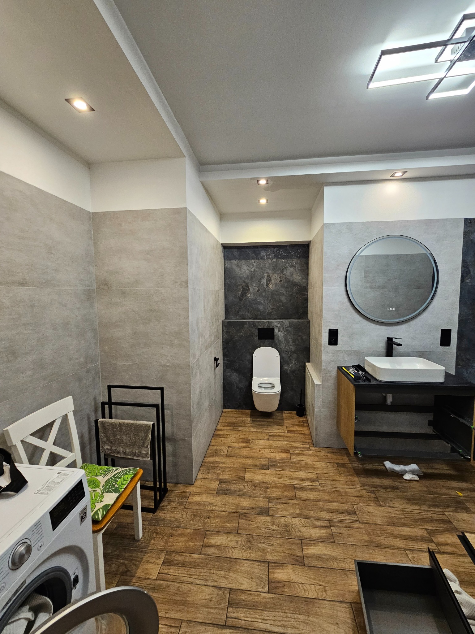 Wykończona łazienka z drewnopodobną podłogą, szarą glazurą, okrągłym lustrem z podświetleniem i pralką. Widoczne detale: grzejnik drabinkowy i nowoczesna armatura.