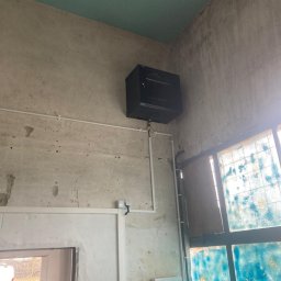 LinkSecure - Czarna szafa rack zamontowana na ścianie z instalacją elektryczną w surowym wnętrzu. Widoczne okno z niebieską szybą i fragment drzwi.