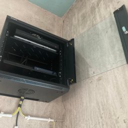 LinkSecure - Otwarta szafa rack Lanberg na ścianie z zamontowanym sprzętem Dahua, widoczne kable. Szara ściana z surowym wykończeniem. Ujęcie z dołu.