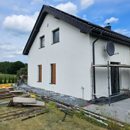 Przem-bud - Biała elewacja domu jednorodzinnego z czarnym dachem, widoczna z boku. Na trawniku leżą materiały budowlane i rusztowanie. Błękitne niebo z chmurami w tle.