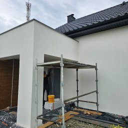 Przem-bud - Biała elewacja domu jednorodzinnego w trakcie prac wykończeniowych, widoczne rusztowanie i pracownik w czarnym ubraniu. W tle maszt telekomunikacyjny.