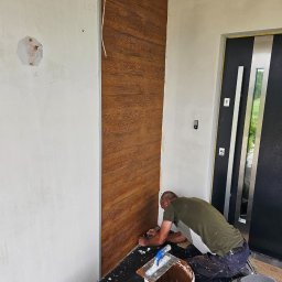 Przem-bud - Pracownik klei panel dekoracyjny imitujący drewno na ścianie przy wejściu do domu. Widoczne narzędzia i wiadro z klejem. Fragment drzwi wejściowych.