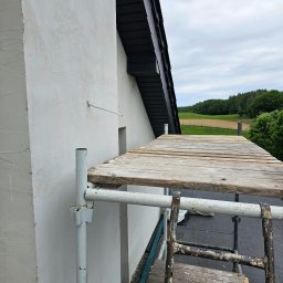 Przem-bud - Ściana budynku po szpachlowaniu, widoczne rusztowanie i drabina. Tło stanowią pola i lasy pod pochmurnym niebem.