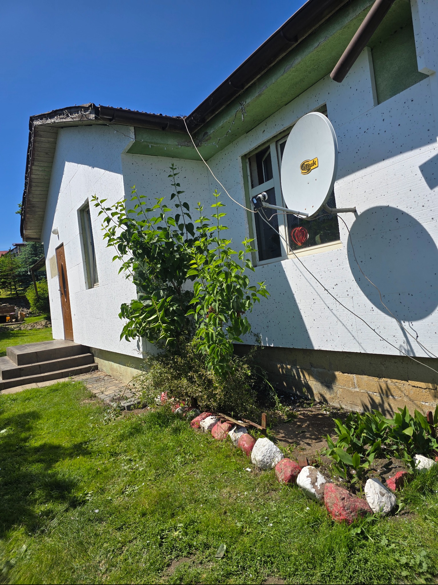 Ocieplony dom z elewacją w trakcie prac, widoczny styropian i siatka. Na ścianie zamontowana antena satelitarna. Ogródek z kamieniami.